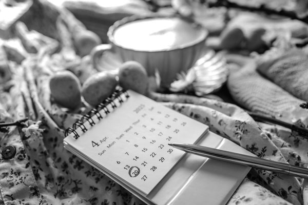 Close-up of an April 2025 calendar with April 20 marked, lying on a floral fabric beside speckled Easter eggs, a cup of coffee, and a golden pen in warm natural light